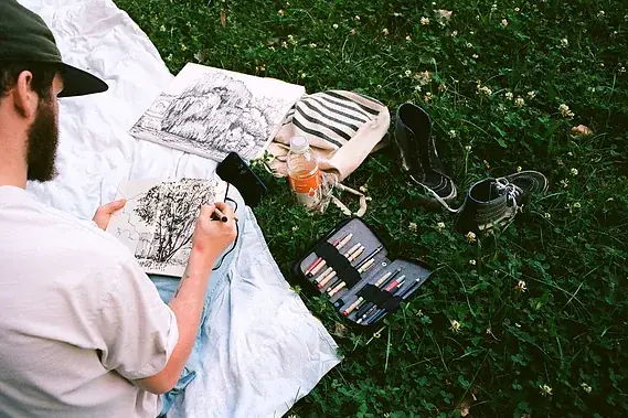 Une personne dessine à l'encre dans un carnet, sa trousse de crayons ouverte à côté d'elle.