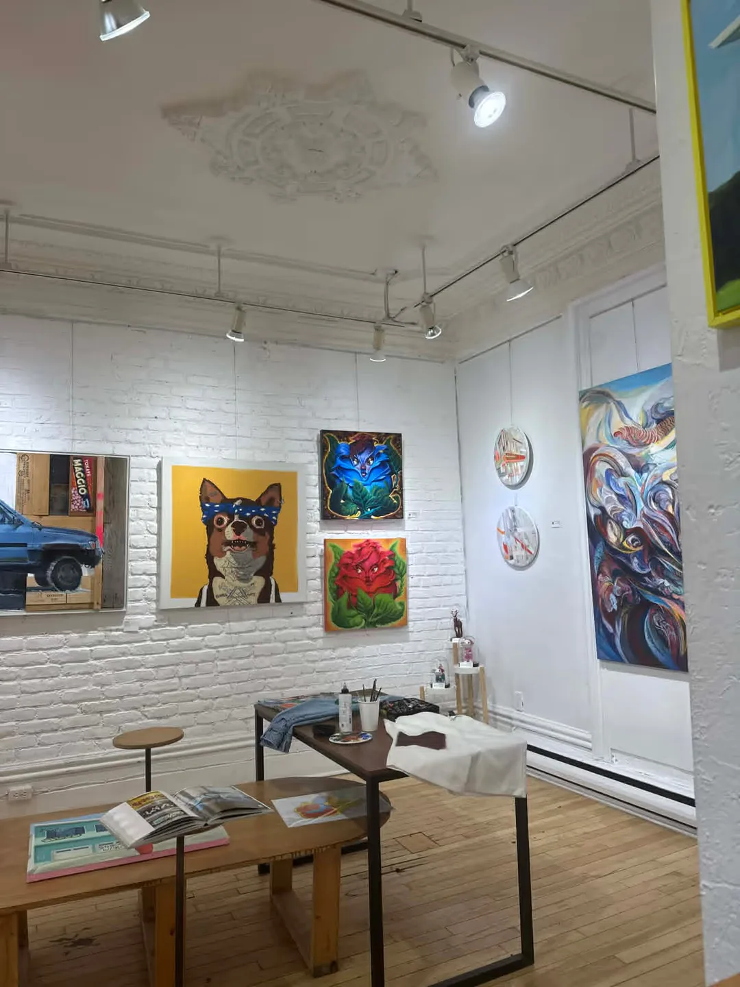 Coin atelier de la galerie avec plafond à moulures, toiles accrochées et table de travail.